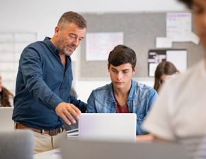 A teacher helps a student using a laptop, highlighting the current teacher shortage.